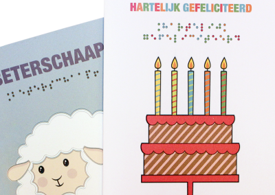 Two greeting cards overlap each other. The card in the back is a light blue greeting card with the text Get Well in large print and braille and below that a relief image of a sheep is printed on the card. The front card features the text CONGRATULATIONS in multicolored black print and Braille, below which is a relief image of a cake with five candles in different colours.