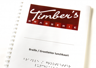 Een boekje met een ringrug met daarop in sierlijke letters de Tekst Timber's Brasserie op een rode achtergrond. Op een witte achtergrond in grote zwarte letters de tekst Braille / Grootletter lunchkaart hieronder staat dezelfde tekst in braille.