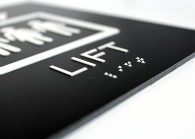 A close-up of a rectangular black DiBond sign. An elevator icon and the letters elevator are printed in relief. Lift is also printed in Braille.