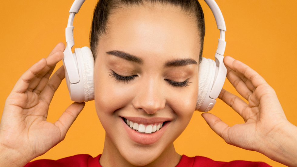 A woman smiles with her eyes closed and she listens to headphones. She has her fingertips on the headphones.